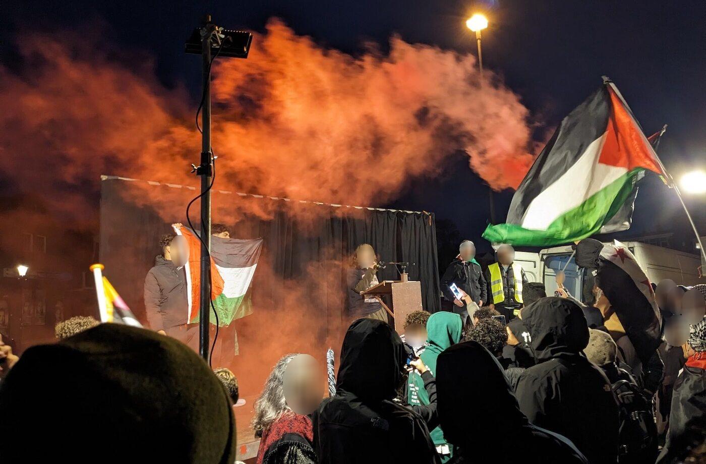 Een grote menigte verzamelt zich in het avondlicht met straatlantaarns rond een podium. Op het geeft iemand een toespraak. Ook staan er verschillende mensen op het podium die prominent een grote Palestijnse vlag omhoog gehouden. Mensen in de menigte dragen voornamelijk jassen en hoodies, wat aangeeft dat het een koele avond is. Een aanzienlijke hoeveelheid oranje-rode rook waait in de lucht en verduistert sommige delen van de scène.