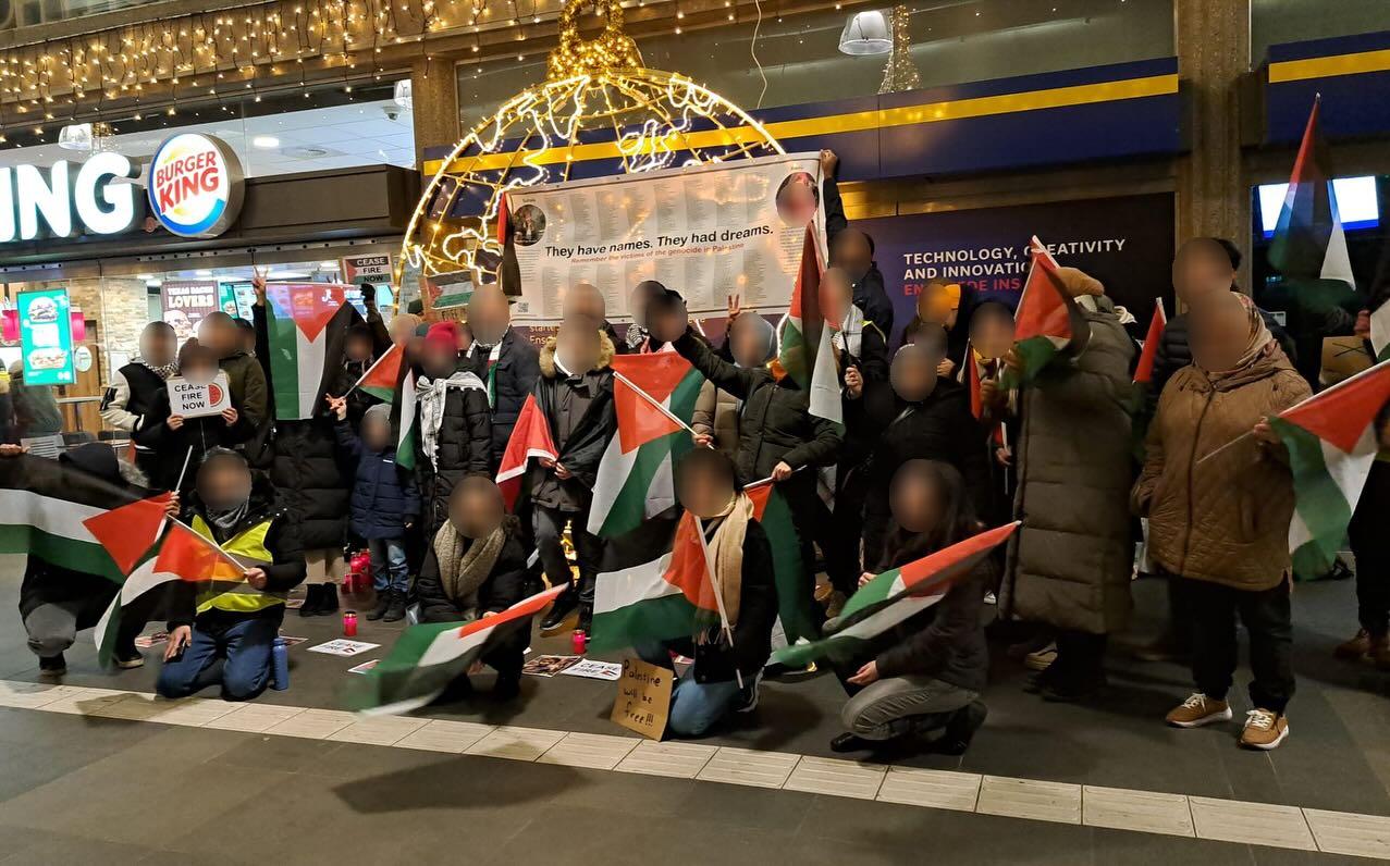 In een verlichte binnenruimte staan en zitten mensen met Palestijnse vlaggen langs een gevel. Verschillende mensen maken met hun vingers het 'V'-symbool voor overwinning. Sommigen hebben borden waarop staat (in het Engels) 'Staakt-het-vuren nu' en 'Palestina zal vrij zijn'. In de achtergrond wordt door verschillende mensen een spandoek met de titel (in het Engels) 'Zij hebben namen. Zij hadden dromen' opgehouden. Op het spandoek staan namen van personen die door de zionistische bezetter vermoord zijn.