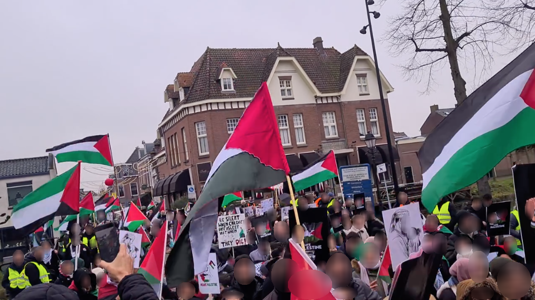 In een historisch stadscentrum lopen op een grauwe dag honderden mensen met Palestijnse vlaggen en borden in solidariteit met Palestina. Borden zeggen onder andere (in het Engels): 'Wees stil wanneer kinderen slapen, niet wanneer ze doodgaan', en borden van een ouder die een dood, gewond of stervend kind omarmt.
