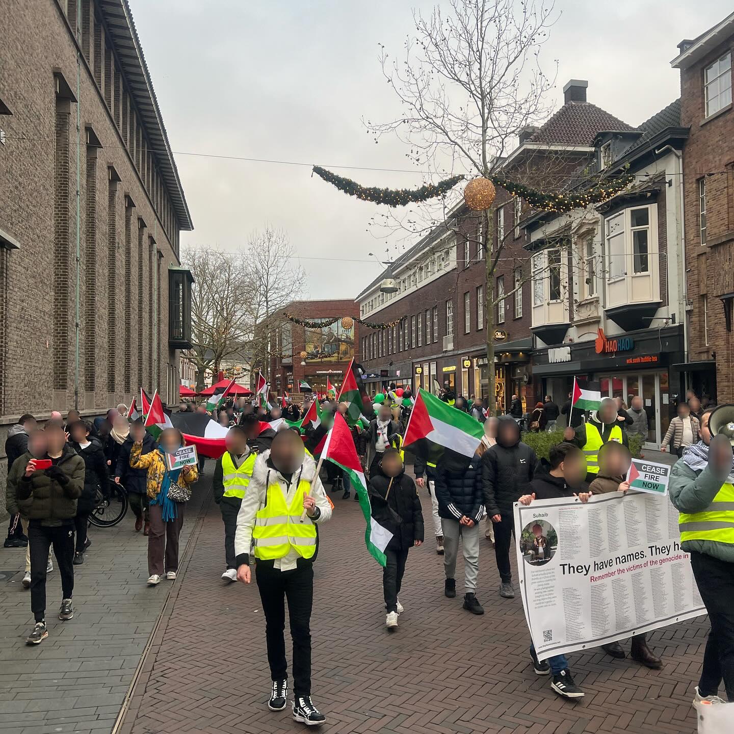 Honderden mensen lopen op een grauwe dag door een winkelstraat waarin winterverlichting hangt. Zij dragen Palestijnse vlaggen en borden met daarop oproepen tot een staakt-het-vuren. Vooraan de stoet rechts in beeld dragen kinderen een groot spandoek met daarop (in het Engels) de tekst 'Ze hebben namen. Ze hadden dromen', met daarbij de namen van duizenden mensen die door de zionisten vermoord zijn.