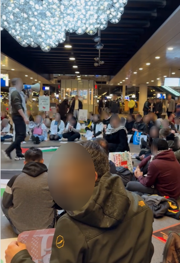 Tientallen mensen zitten in een cirkel op de grond met spandoeken en borden. Eén persoon staat in het midden van de cirkel met een megafoon.