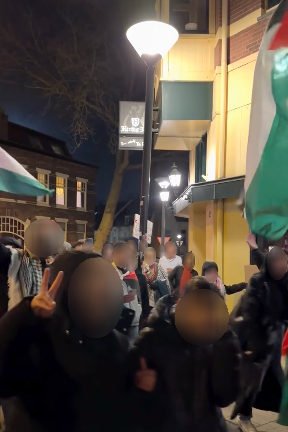 Een stoet mensen met Palestijnse vlaggen marcheert bij nacht en straatverlichting. Een persoon op de voorgrond maakt een V-teken ter overwinning met diens vingers.