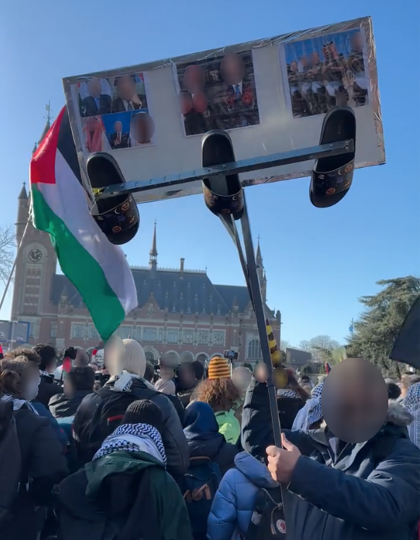 Mensen staan op een open plaats voor een gebouw in traditionele architectuur. Zij houden borden en vlaggen op. Op de voorgrond staat een persoon met een bord met daarop portretten van verschillende oorlogsmisdadigers, waar met een mechaniek een rij van drie schoenen tegenaan wordt geklapt.