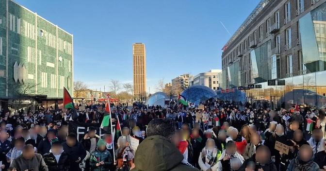 Honderden zo niet duizenden mensen vullen een plein op een heldere dag. Zij luisteren naar een spreker en dragen Palestijnse vlaggen en borden.