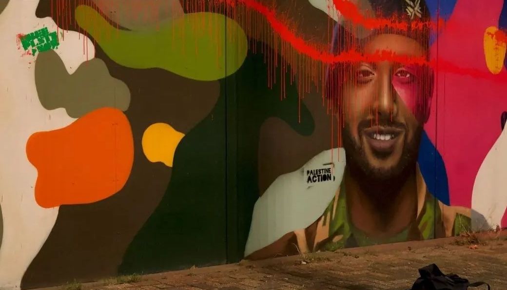 Een muurschildering aan de onderkant van een hek met daarop kleurrijke legerpatronen en een lachend persoon in militair uniform. Over de muurschildering is op meerdere plaatsen rode verf gespoten, plus teksten (in het Engels) zoals: 'Bevrijd Palestina nu' en 'Palestina Actie'. Op de afbeelding zijn met witte en rode letters op een zwarte achtergrond digitaal teksten geplaatst, in Nederlands en Engels: 'Geen steun voor genocides (...) betekent geen dienstplicht', plus het logo van de groep Palestine Action.