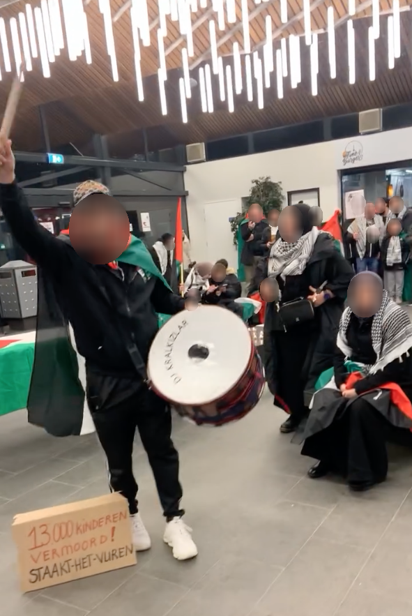 Mensen staan en zitten in een goedverlichtte stationshal. Sommigen hebben Palestijnse vlaggen om en dragen keffiyeh. Een persoon bespeelt een trommel.