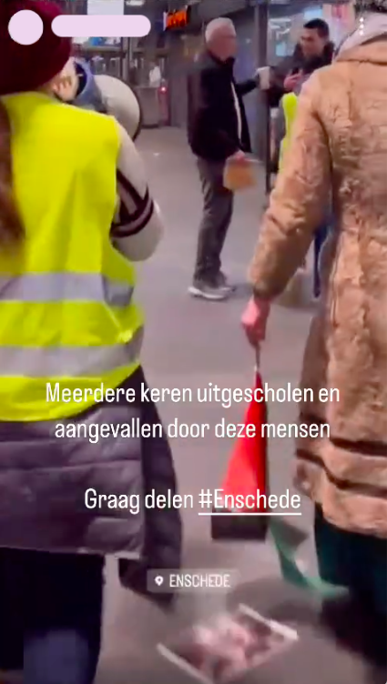 Een kil verlichtte stationshal waar twee personen op een afstandje van voren te zien zijn, en twee personen dichtbij op de rug. Eén persoon die op de rug te zien is heeft een megafoon in de hand, de ander houdt een Palestijnse vlag vast. Op de grond ligt tenminste één flyer. Over het beeld heen staat in witte letters: 'Meerdere keren uitgescholden en aangevallen door deze mensen. Graag delen #Enschede', met daaronder nogmaals een locatie-icon en het woord 'Enschede'.