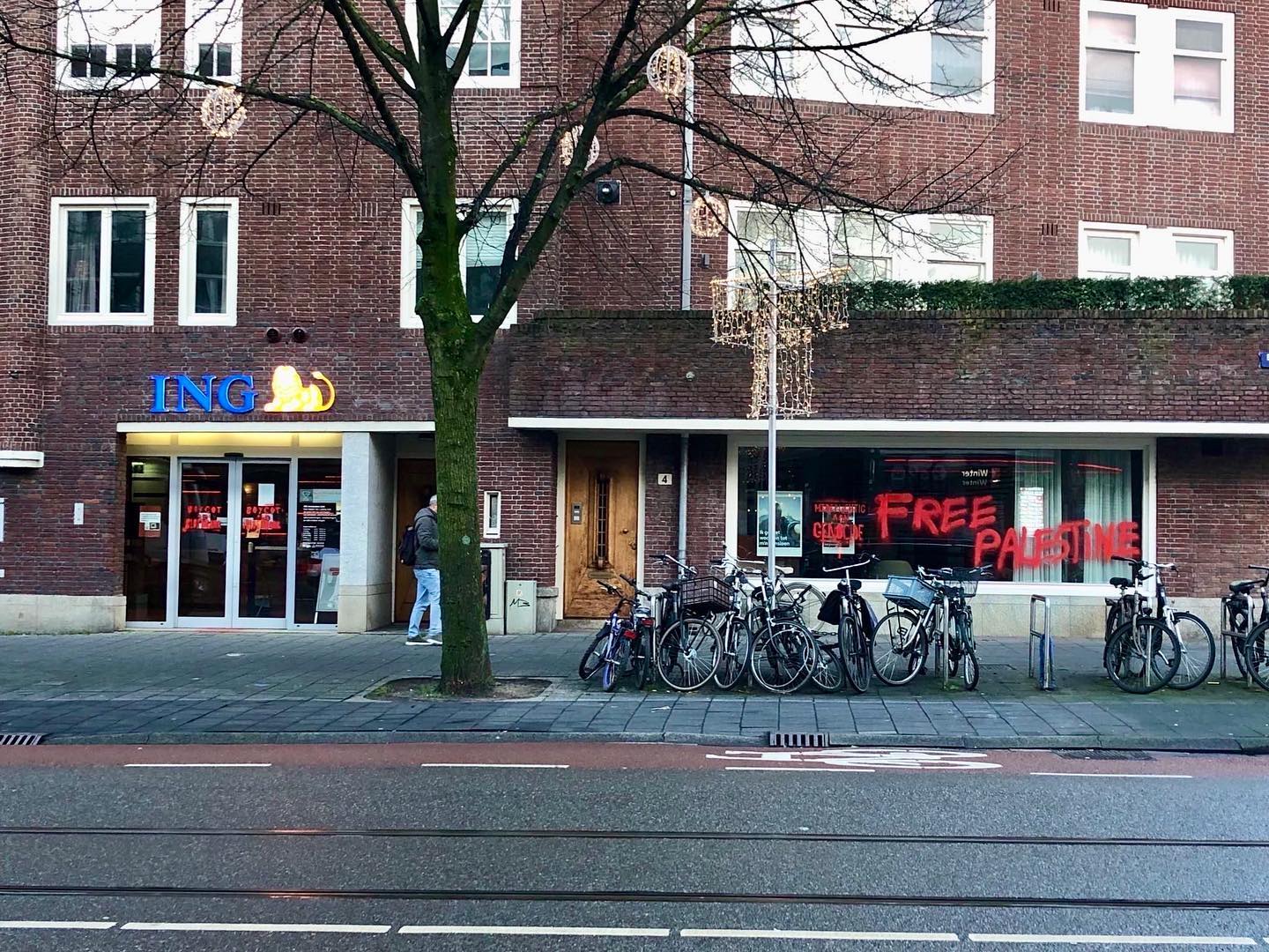 Een straataanzicht op een ING bankfiliaal op een begane grond, met boven het filiaal appartementen. Voor het filiaal staan stallingen met fietsen. Op de schuifdeuren staat in rode verf de tekst 'Boycot deze bloedbank'. Op een raam een stuk aan de rechterkant staat de tekst 'Medeplichtig aan genocide' en (in het Engels) 'Bevrijd Palestina'.