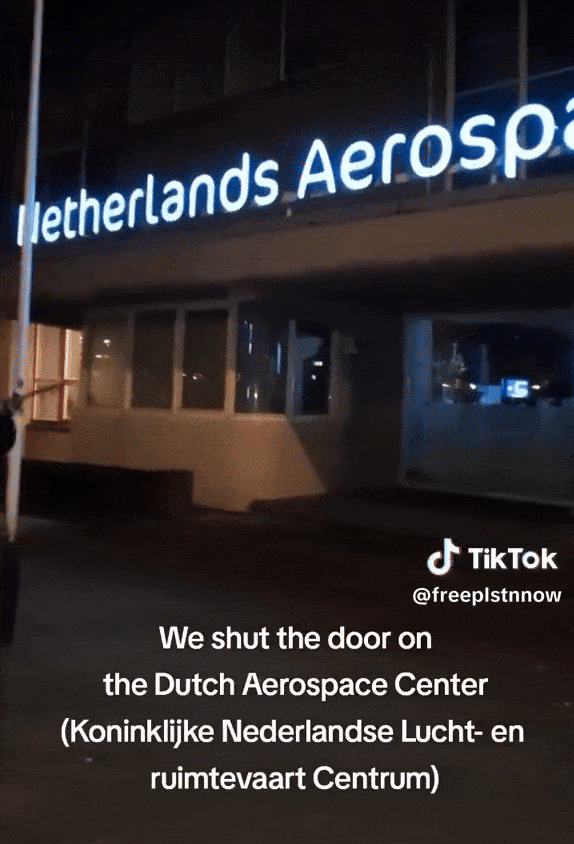 'Een vooraanzicht, in het donker op de geval van een gebouw van het Koninklijke Nederlandse Lucht- en Ruimtevaartcentrum in Amsterdam. Een persoon bespuit de gevel met rode verf.'