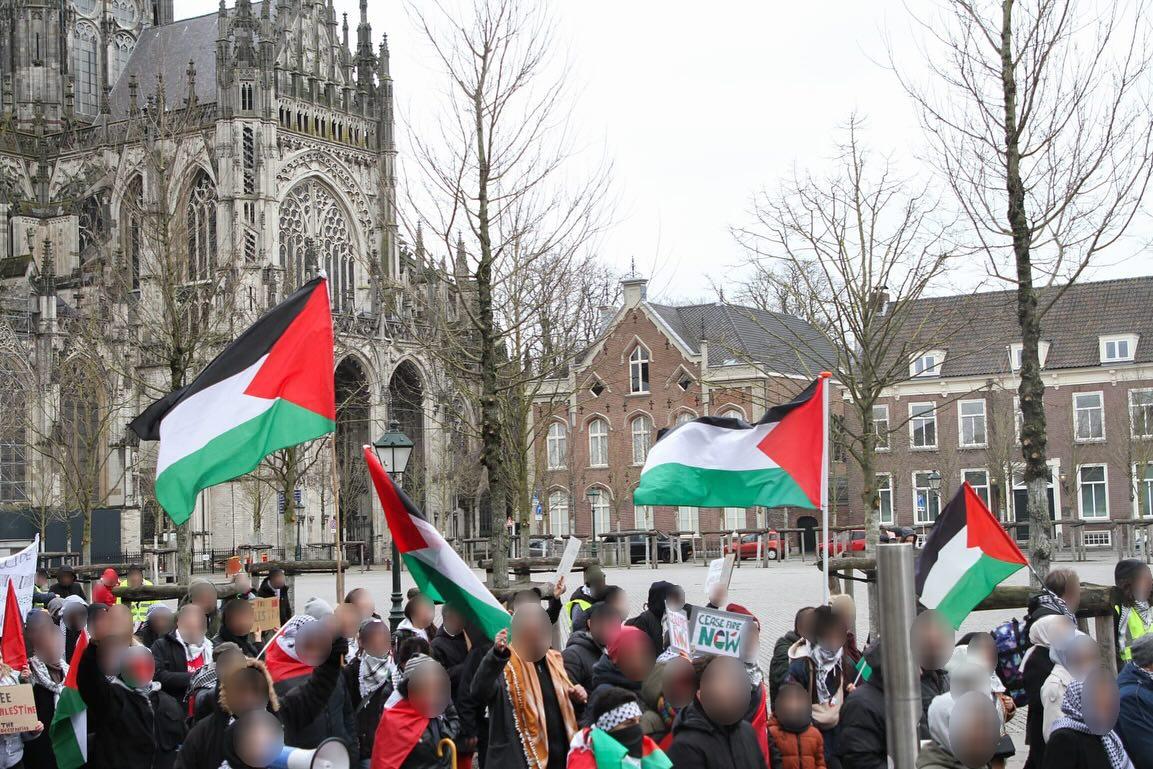 Een grote groep mensen loopt langs een zijde van een open plein met Palestijnse vlagge, borden, megafoons en kuffiya. Op de achtergrond de ruimte van het open plein met enkele statige gebouwen en een gigantisch gotisch kerkgebouw.
