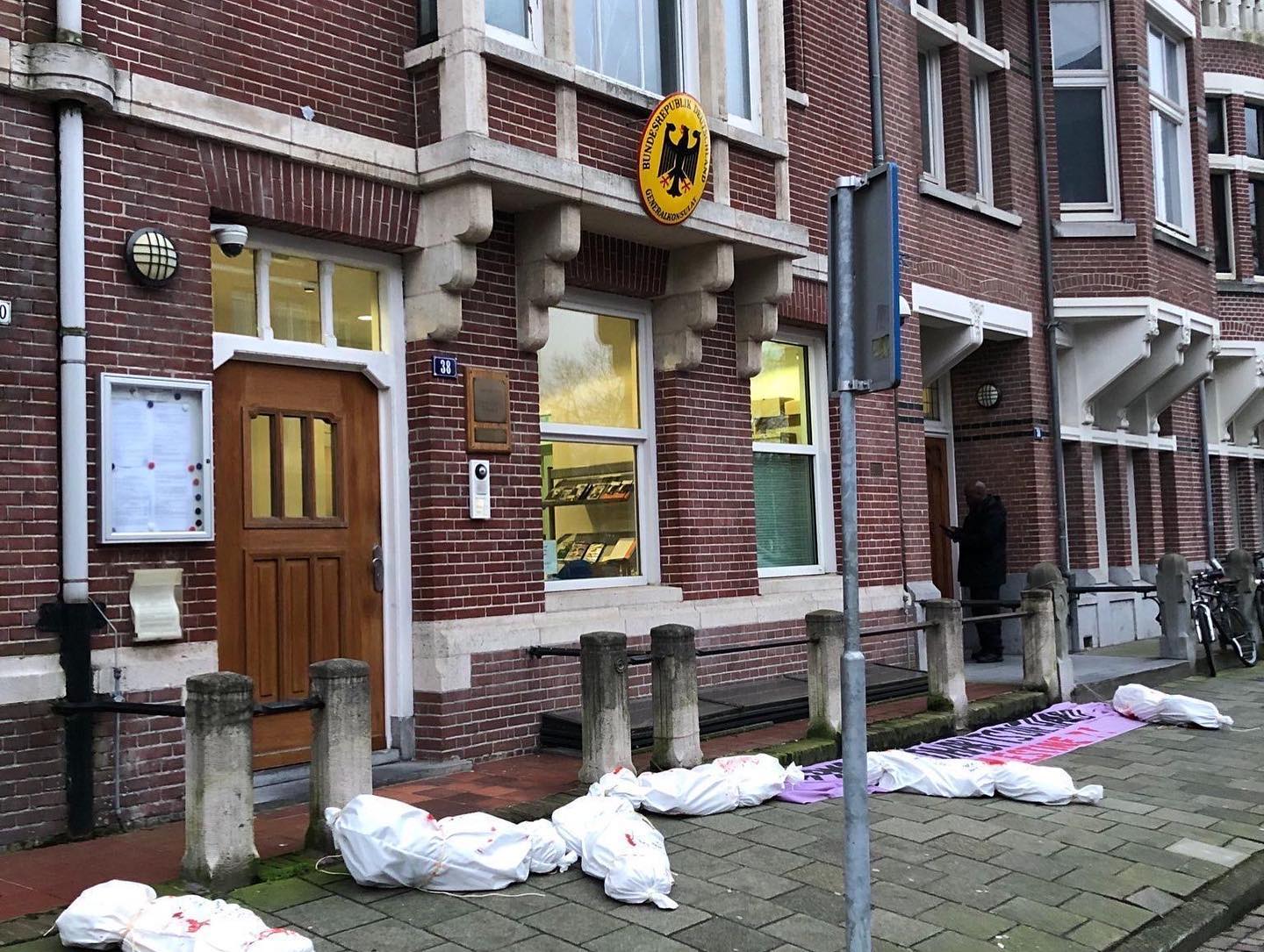 Aanzicht op het Duitse consulaat in Amsterdam. Op de stoep voor het gebouw liggen verschillende figuren die de indruk geven van bloedige lichamen in lijkzakken gewikkeld. Ook ligt er op de grond een spandoek met de tekst (in het Engels) 'Duitsland: oude gewoontes raak je maar lastig kwijt. Bevrijd Palestina'.