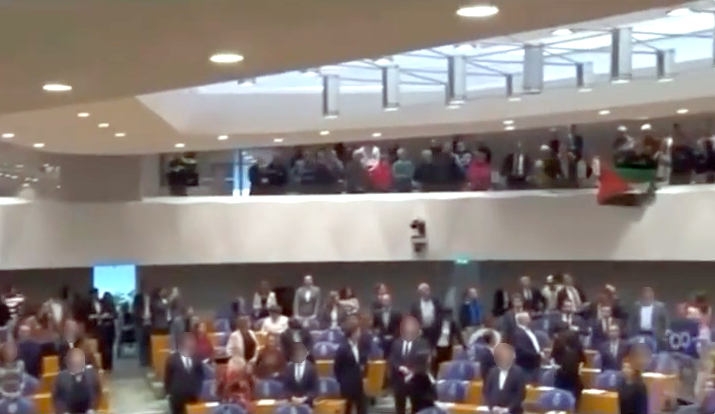 Een zicht op een open zaal met een oplopende tribute met blauwe en houten zitgedeeltes, waarin op de meeste plaatsen mensen staan. Op een balustrade op de eerste verdieping staan ook mensen. Eén van hen houdt een Palestijnse vlag over de rand van de balustrade.