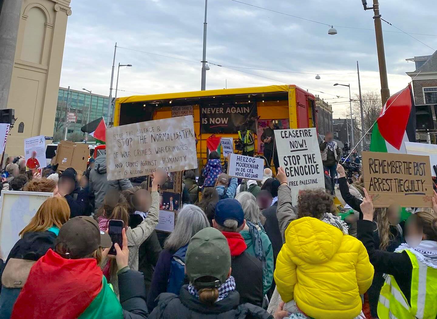 Op een grauwe dag staan op een plein mensen met borden en Palestijnse vlaggen met hun gezichten gericht naar een gele, overdekte, open kar. Achterin de kar hangen spandoeken. Borden en spandoeken zeggen onder meer (in Nederlands en Engels): 'Dat nooit meer is nu', 'Herzog moordenaar, in de cel voor 100 jaar', 'Staakt-het-vuren-nu, stop de genocide en bevrijd Palestina', 'Onthoudt het verleden, arresteer de schuldigen', 'Stop de stilte & stop het normaliseren van oorlogsmisdagen, oorlogsmisdadigers en misdaden tegen de mensenlijkheid, het doodt iedere dag mensen!'