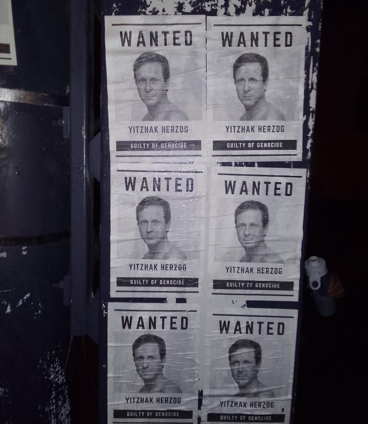 'Een muurgedeelte in de schemering, met daarop een vak van zes posters met daarop een portret van Yitzhak Herzog en de tekst (in het Engels) 'Yitzhak Herzog schuldig aan genocide'. Aan de rechterzijde is een hand met daarin een drinkfles zichtbaar.'