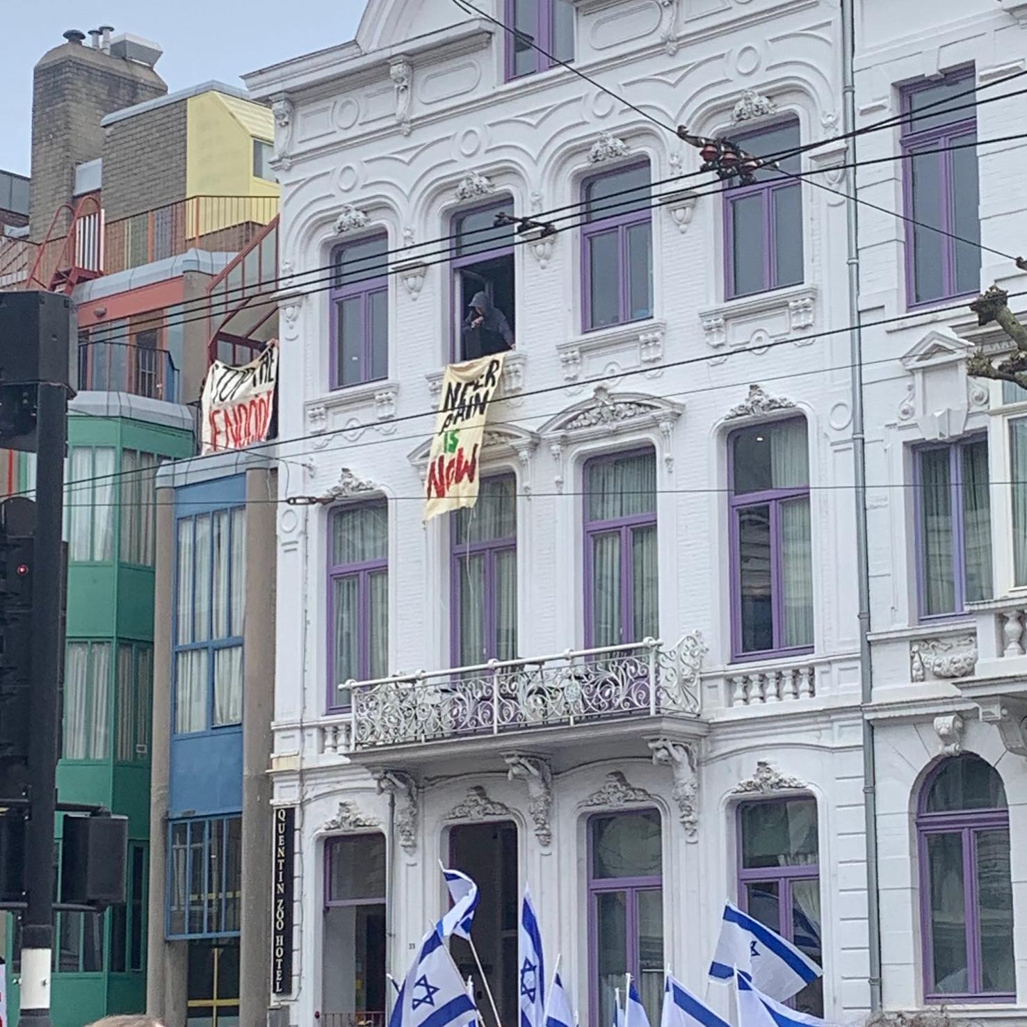 'Vanaf de tweede verdieping van een klassiek hotelgebouw met witte gevel aan paarse kozijnen wordt een spandoek uit het raam gehouden, met daarop de tekst in zwarte, groene en rode letters (in het Engels): 'Dat nooit meer is nu'. Vanuit het naastliggende gebouw in kleurrijke jaren '90 architectuur wordt ook een spandoek opgehouden, met de tekst (in het Engels) 'Stop de genocide'. Onderaan het beeld, op straatniveau, wordt elf vlaggen opgehouden van de zionistische entiteit.'