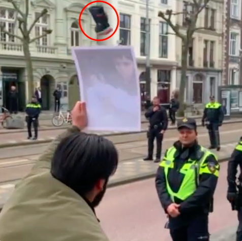 'Blik op een open straat met hoge gebouwen en kale bomen. De straat is bezwaaid met politieagenten, zowel in uniform as in burger. Er staan enkele fietsen geparkeerd, en op de achtergrond is een tramhalte. Op de voorgrond houdt een persoon met een beige jas en donker haar houdt een geprinte foto op richting de agenten. Helemaal bovenin beeld vliegt een halfhoge blauwe laars met lichte zool. De laars is digitaal uitgelicht met een rode cirkel.'
