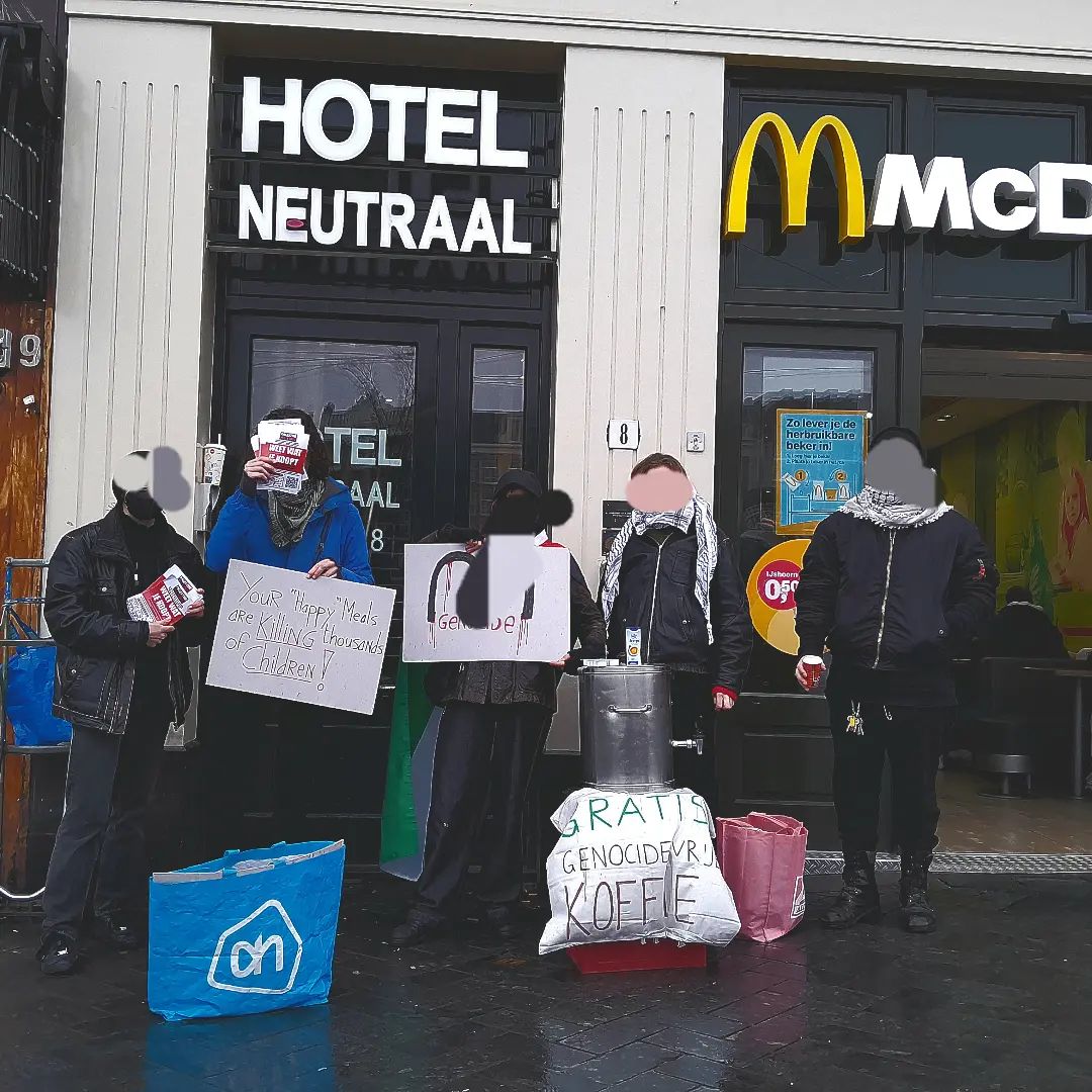 'Een groep van ongeveer vijf mensen in donkere kleding en keffiyeh staat op een regenachtige straat voor de gevel van een filiaal van McDonald's, naast hotel Neutraal. De mensen hebben tassen en flyers bij zich. Ook staat er een groot metalen vat. Twee personen hebben borden in hun handen. Op het ene bord staat (in het Engels): 'Jouw 'Blije Maaltijden' doden duizenden kinderen!', en op het andere bord staat een grote 'M' van het McDonald's logo, met daaronder het woord 'genocide'. De hoofden van de personen zijn digitaal onherkenbaar gemaakt.'