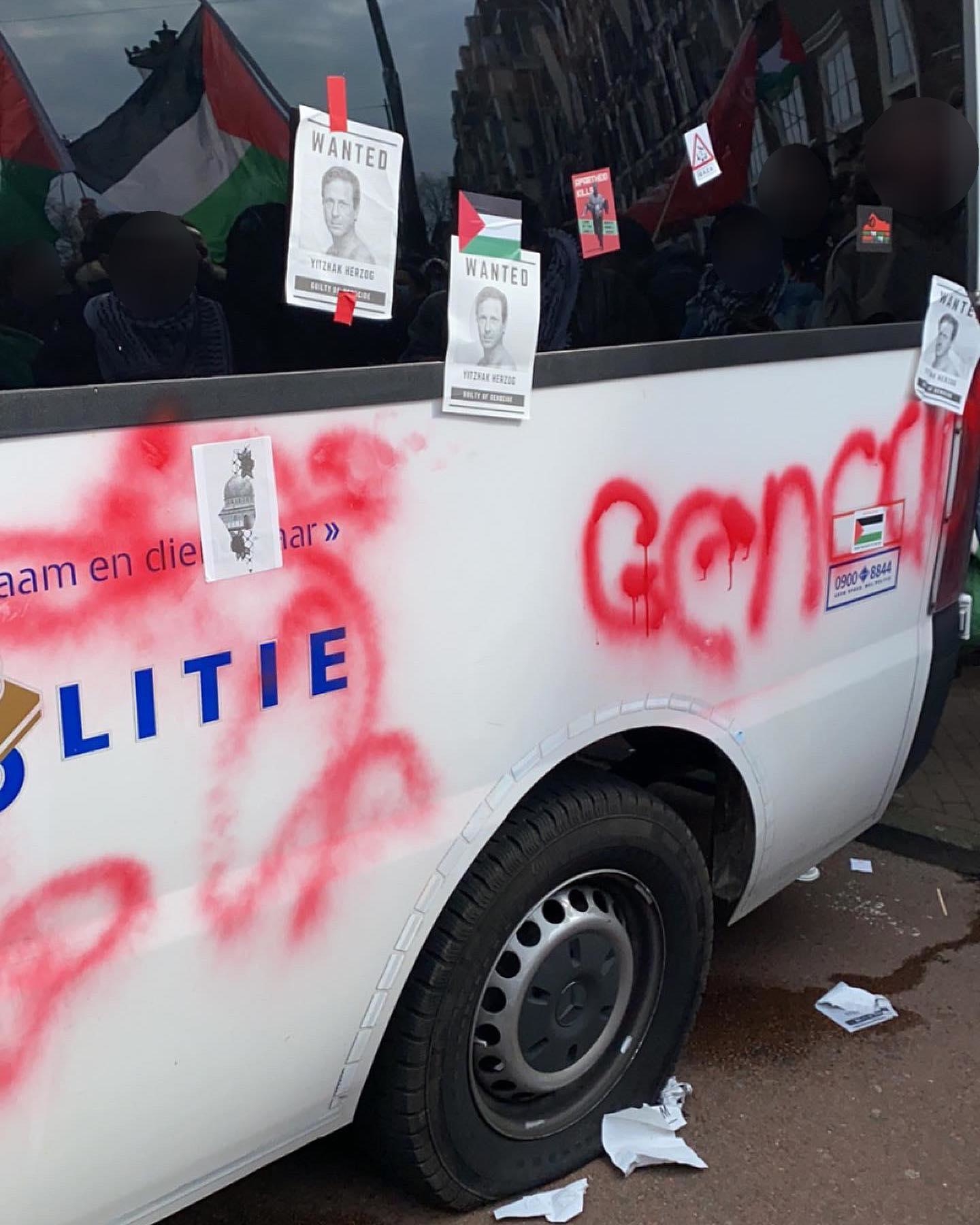 'Het achterste deel van een wit politiebusjs met daarop nog een deel van de tekst 'Politie' en 'Waakzaam en dienstbaar'. Op het busjs zitten stickers die solidariteit uitdrukken met Palestina, en briefjes met daarop het portret van Yitzhak Herzog en de tekst (in het Engels): 'Gezocht'. Ook is onder meer het woord 'genocide' op het busje gespoten. De band van het busje is lek en plat. In de raamweerspiegeling zijn meerdere mensen met Palestijnse vlaggen te zien.'