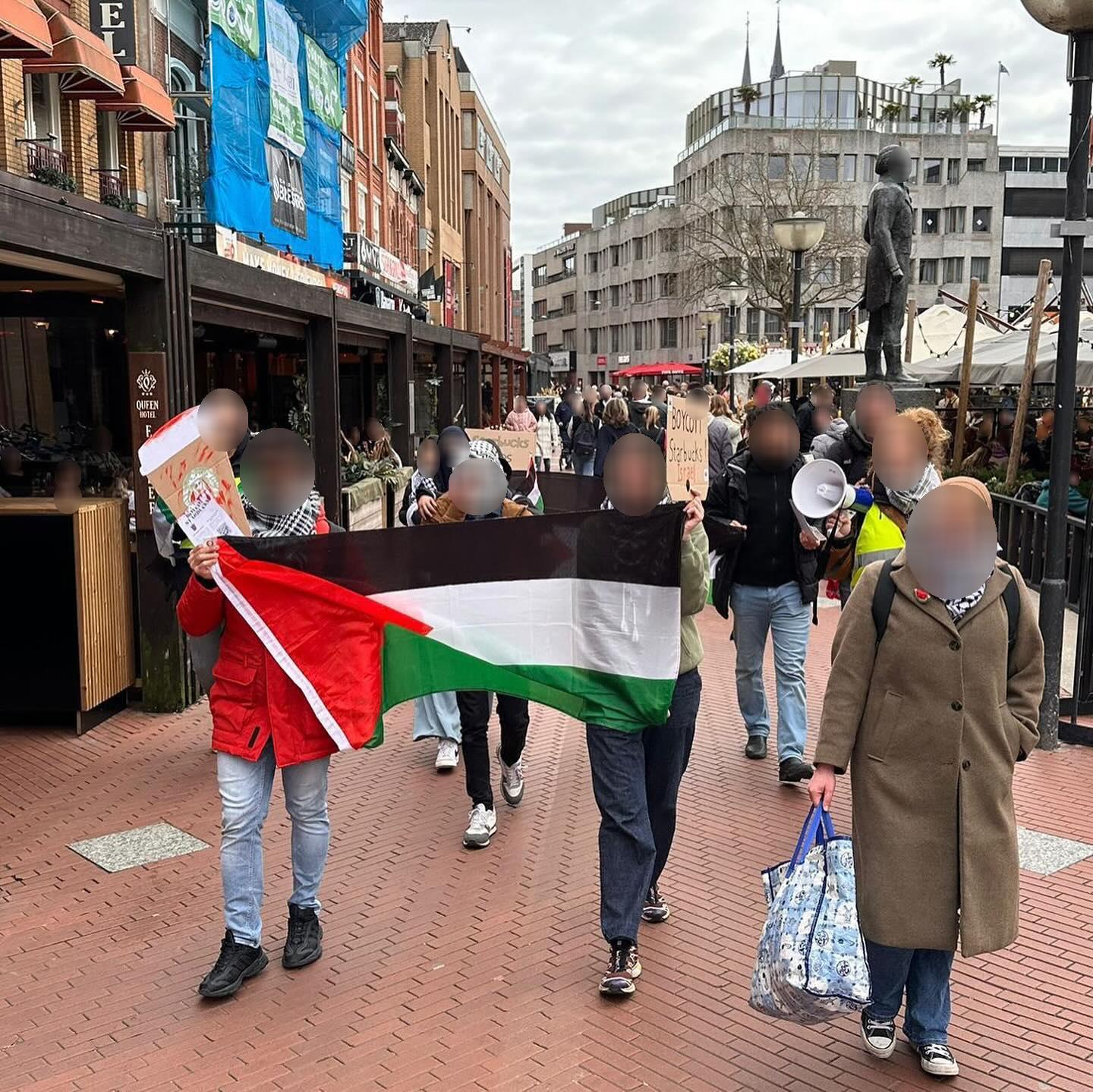 Mensen lopen door een stadscentrum langs terrassen en met op de achtergrond hoge gebouwen en een bronzen standbeeld op sokkel. De mensen dragen Palestijnse vlaggen en borden met teksten zoals 'Boycot Starbucks', of een bord in de vorm van een Starbucks-drinkbeter met bloedspatters erop.