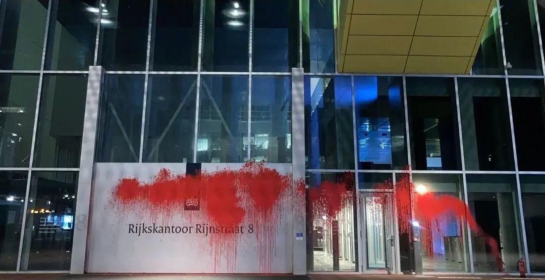 'Aanzicht op de voorgevel van een hoog, glazen gebouw. Een groot grijs bord toont het adres van het gebouw: 'Rijkskantoor Rijnstraat 8', met iets aan de rechterkant een deur. Flinke delen van de voorgevel zijn bespoten met heldere rode verf.'