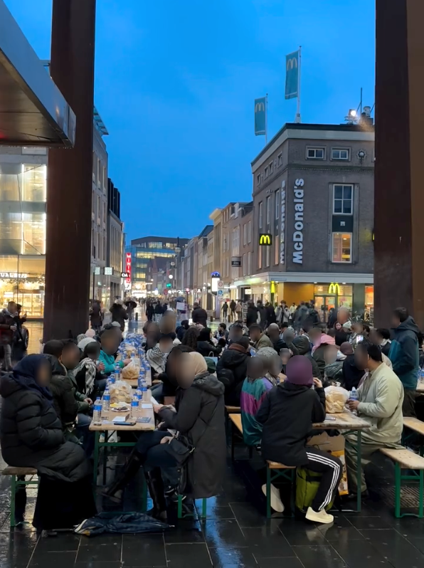Tientallen mensen zitten in de schemer van de avond aan lange tafels in de openbare ruimte. Op de tafels staat eten en drinken uitgestald. Op de achtergrond een winkelstraat.