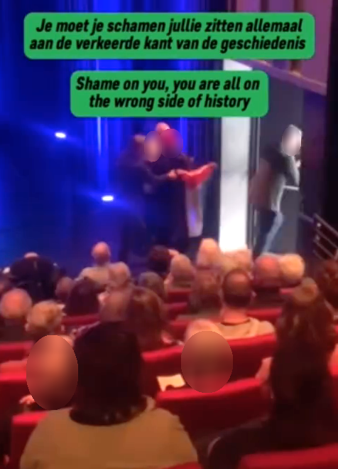 In een donkere concertzaal met oplopende tribune wordt een persoon met een Palestijnse vlag van een podiumvloer weggevoerd.