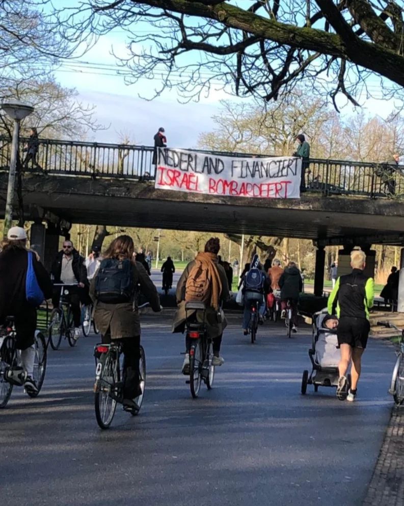 'Aanzicht op een gemengd wandel- en fietspad in een parkachtige omgeving met bomen en gras, waarop zo'n twintig mensen zichtbaar zijn. Dwars boven het pad loopt een brug. Vanaf de brug laten twee personen een wit spandoek met daarop in rood-zwarte letters de boodschap: 'Nederland financiert, Israël bombardeert'.'