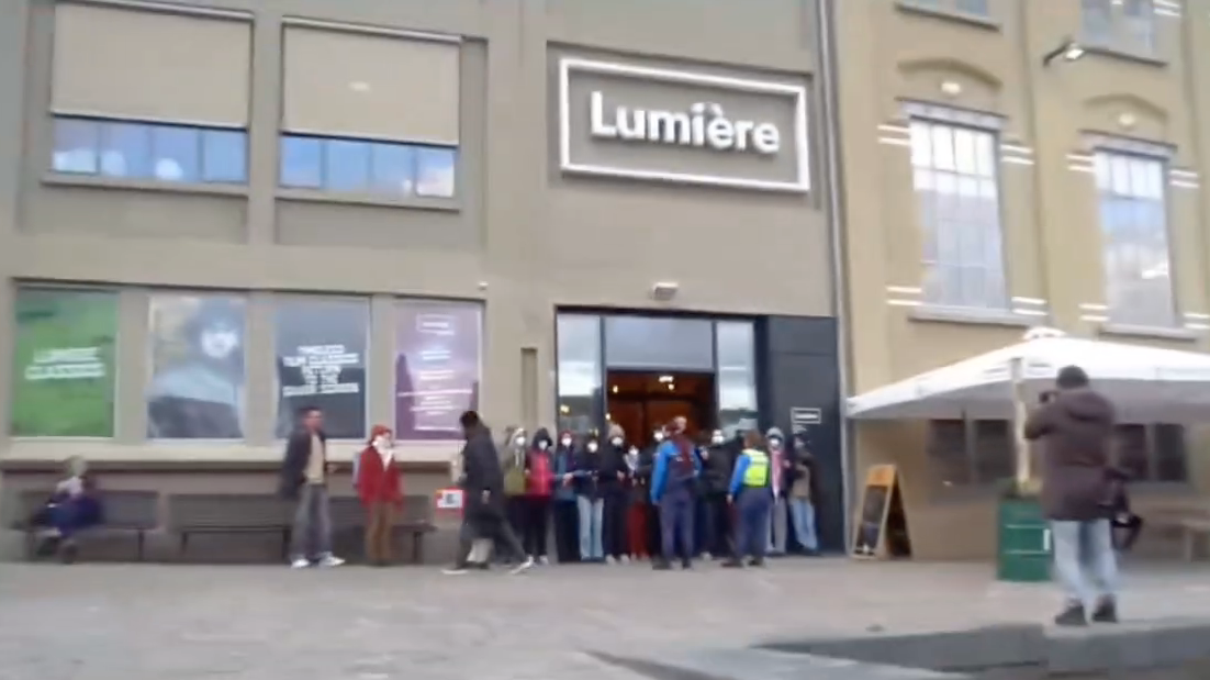 Aanzicht op een plein met een hoog gebouw in grauw-geel steen, hoge verdiepingen en hoge ramen. Voor het gebouw staat een terrasparasol. In het midden van het beeld staat boven de ingang in grote, witte letters het woord 'Lumière'. De ingang zelf is een hoge, donkere duur. Voor de ingang staan zo'n twintig mensen met de armen ineen gehaakt, hun gezicht richting het plein. De meesten van hen dragen witte gezichtsmaskers. Voor hen staan twee handhavers in uniform. Drie personen lopen langs, een ander maakt verderop een foto van de situatie, en weer een ander zit op een bankje.