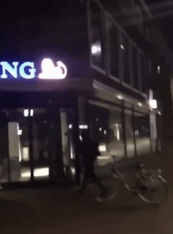 Een persoon komt midden in de nacht op de fiets aanrijden bij een filiaal van de ING bank. Voor de ingang stapt de persoon af, pakt een apparaat, en begint de voorgevel van het filiaal en het lichtgevende logo van de ING rood te spuiten.
