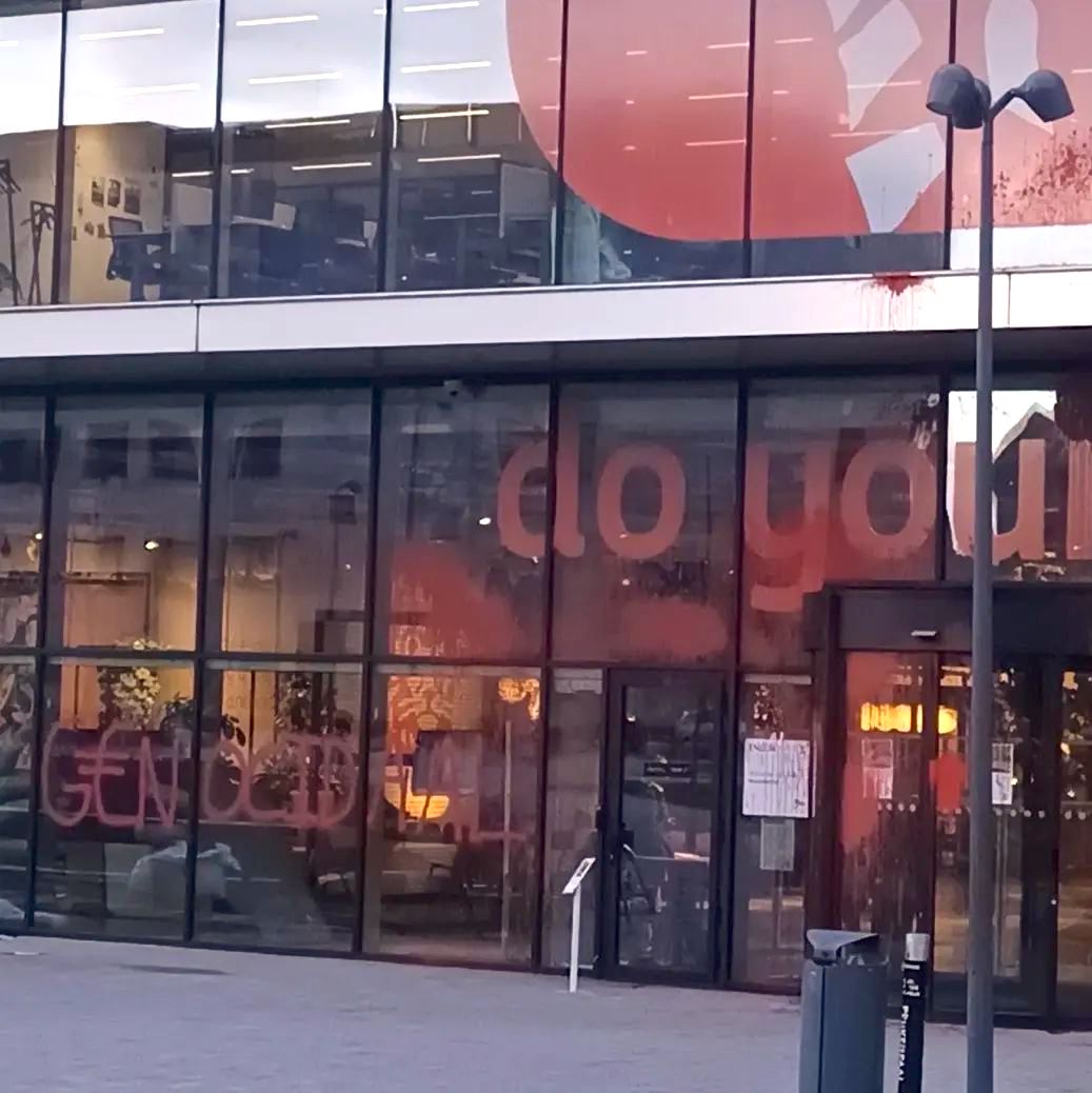 De glazen voorgevel van een ING-kantoor, met meerdere verdiepingen met wachtruimte, kantoren en bureaus van buitenaf zichtbaar. Op de voorgevel staat ook een leeuw van het ING-logo en de tekst (in het Engels) 'doe je ding'. Op de ingangdraaideur en meerdere delen van de gevel zitten rode vlekken verf. Ook staat er het woord 'genociaal' gespoten.