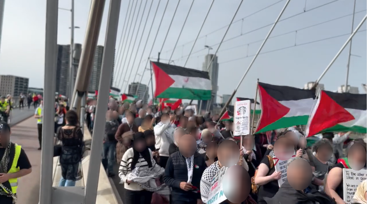 Duizenden mensen Palestijnse vlaggen en borden marcheren op een heldere dag over de autobaan een brede, lange brug. Op het fietspad aan de linkerzijde lopen mensen in fluoriscerende gele hesjes en staat politie op de fiets. Eén van de borden roept mensen op Starbucks te boycotten.