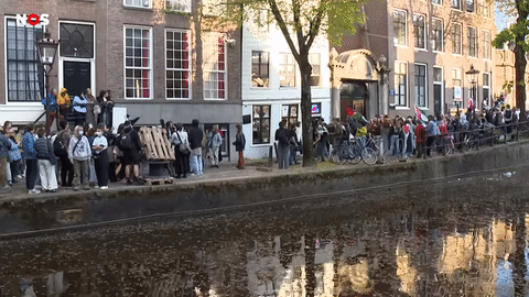 Een kort bewegend fragment aan een gracht, waarin tientallen zo niet honderden mensen in een keten hout en andere objecten bijeenbrengen om een barricade te versterken. Tientallen andere mensen kijken toe vanaf de andere kant van de barricade. Het is een zonnige dag.