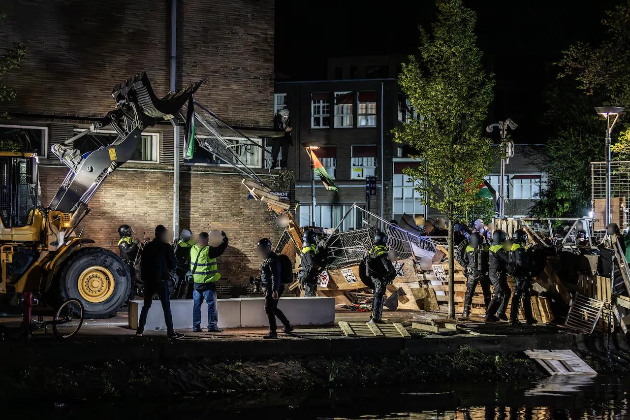 Een nachtelijke straatscène. Een zware gele graafmachine staat prominent op de voorgrond, schuin naar het midden gericht. Meerdere mensen, gekleed in donkere kleding, waaronder politieagenten in donkere uniformen, sommigen met fluorescerende gele veiligheidsvesten, zijn zichtbaar bij de bulldozer en verspreid rond een tijdelijke barricade bestaande uit houten planken en barrières. De tijdelijke constructie strekt zich gedeeltelijk uit tot en rond een water. Meerdere verlichtte gebouwen, veelal van baksteen, zijn zichtbaar op de achtergrond, net als wat bomen. Op de voorgrond bevinden zich zo rond de vijftien politieagenten met knuppels die tegen de barricade en dranghekken aanduwen. Achter de barricade, en op een verhoging, staan een stuk of twintig demonstranten die veelal keffiyeh dragen en een enkele Palestijnse vlag hooghouden.