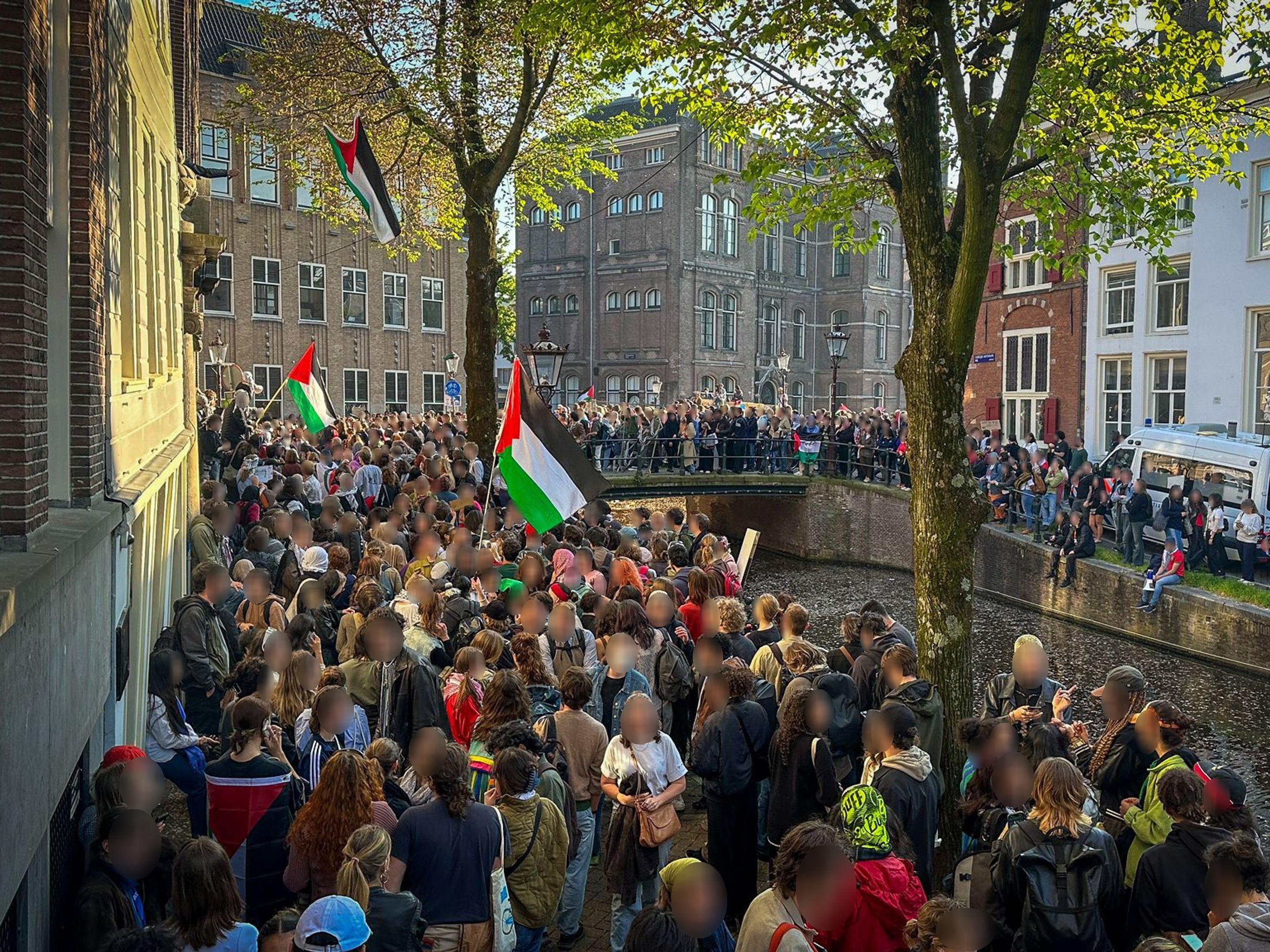 Een grote, dichte mensenmassa protesteert in een stadsstraat aan beide zijden van een gracht. De menigte draagt borden en zwaait met Palestijnse vlaggen.