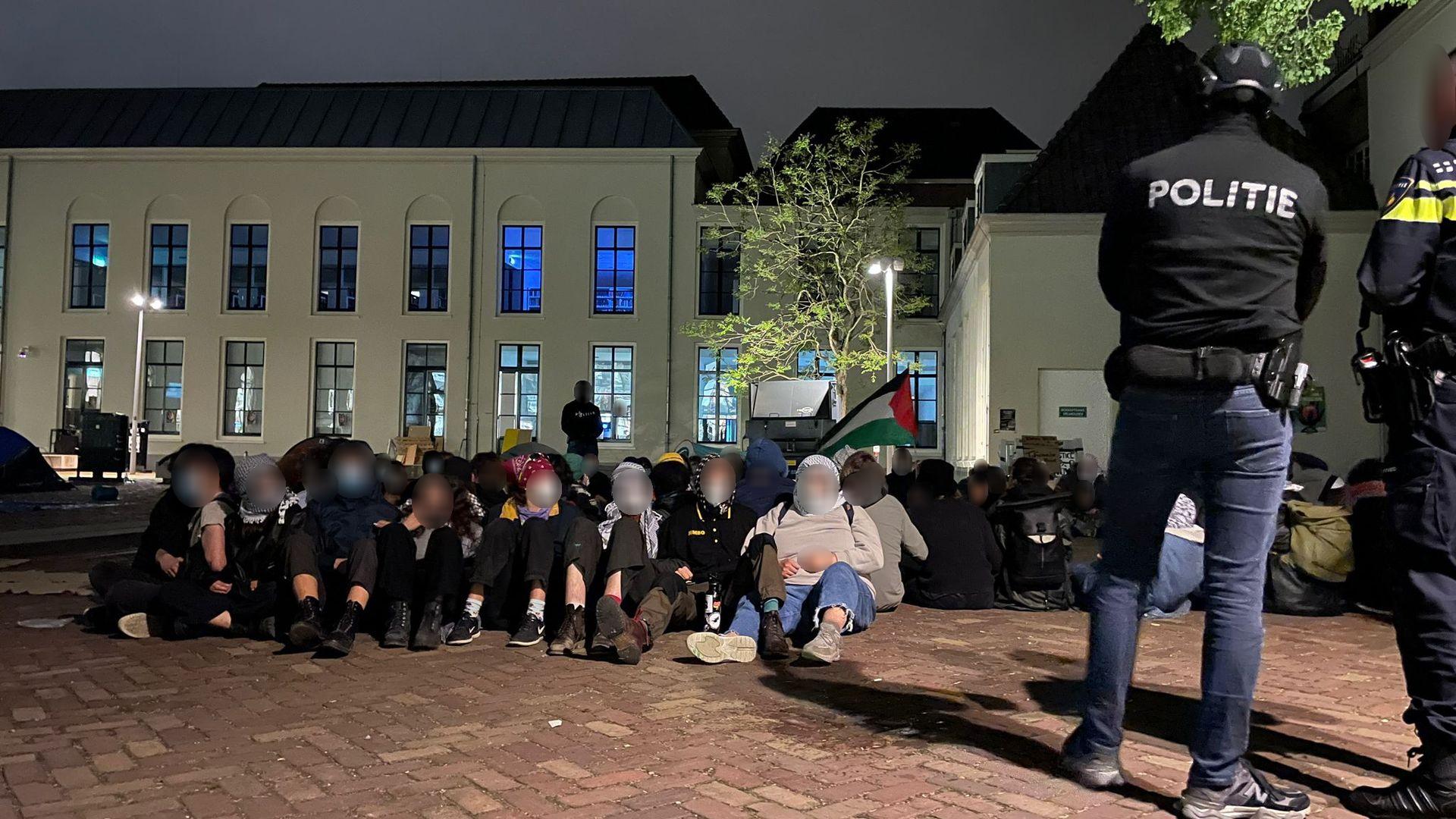 Een nachtelijk protest. Een groep van ongeveer vijftig mensen zit op een geplaveide, bakstenenachtige grond. Velen dragen maskers. In het midden van de groep zwaait iemand met een Palestijnse vlag. Achter hen is een groot, lichtbeige gebouw zichtbaar met meerdere hoge ramen, en een door straatlantaarns verlichtte boom. Er zijn tenminste drie politieagenten in uniform met maskers, helmen, wapens en handboeien zichtbaar. Eén van hen is van op de rug zichtbaar met daarop groot de letters 'POLITIE'.
