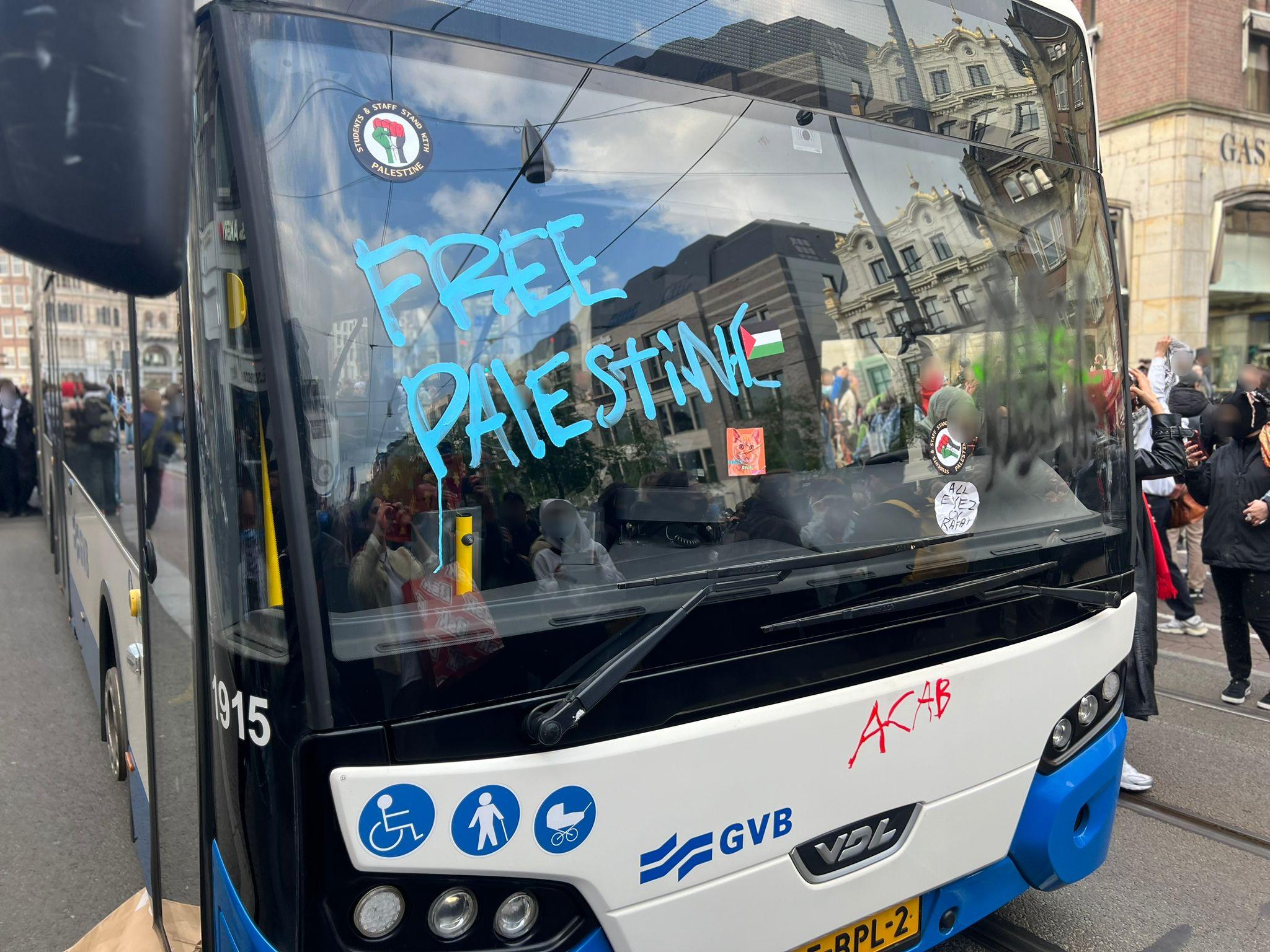Een wit-blauwe bus van de Amsterdamse vervoerder GVB staat midden op de weg stil, met op de voorruit en voorkant verschillende protestboodschappen. Onder meer staat er in grote blauwe stiftletters 'FREE PALESTINE' ('BEVRIJD PALESTINA'), stickers met een Palestijnse vlag erop, en met rode stift de afkorting 'ACAB' ('Alle politieagenten zijn schurken'). Verschillende mensen staan om de bus heen, waarvan sommigen maskers en keffiyeh dragen.