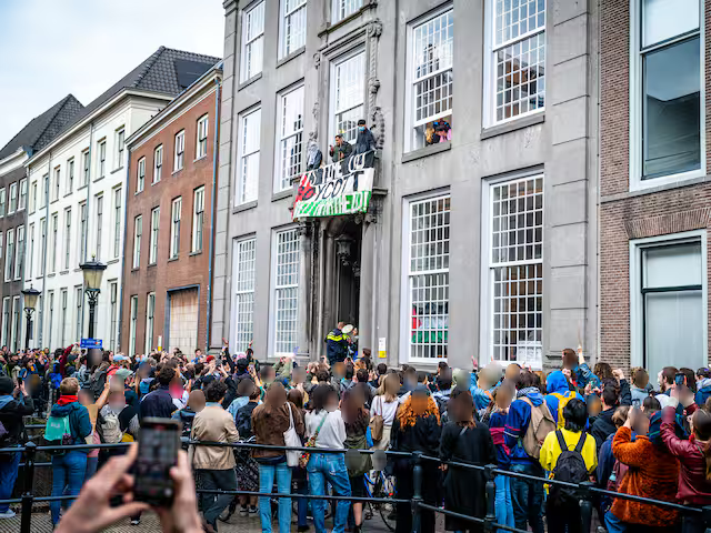 Een grote menigte mensen heeft zich verzameld voor een rij historische gebouwen met hoge, bakstenen gevels en grote ramen. De menigte kijkt naar een aantal personen dat op een balkon op de eerste verdieping van één van de gebouwen staat. Vanaf het balkon hangt een grote banner, mogelijk met de boodschap 'Het is tijd dat UU apartheid boycot' (de tekst is niet volledig zichtbaar). Meerdere mensen in de menigte houden hun telefoons omhoog, mogelijk om de gebeurtenis te fotograferen or filmen. Een metalen reling, mogelijk van een brug, scheidt de menigte van andere delen van de straat. Het is gedeeltelijk bewolkt.