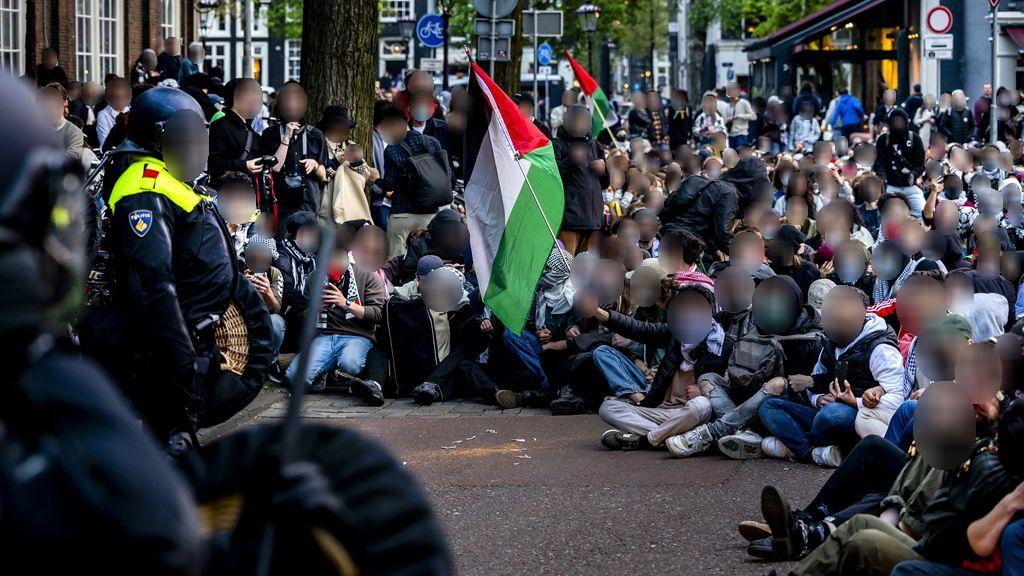 Een groep van duizenden demonstranten zit op een plein in een dichte, georganiseerde opstelling. Velen dragen keffiyeh en gezichtsmaskers. Een grote Palestijnse vlag wordt prominent omhoog gehouden door een van de demonstranten. Aan weerszijden van de demonstranten staat een rij politieagenten in donkere uniformen staat. Het is droog. Op de achtergrond zijn een café, trottoirs en een groene buitenomgeving zichtbaar.
