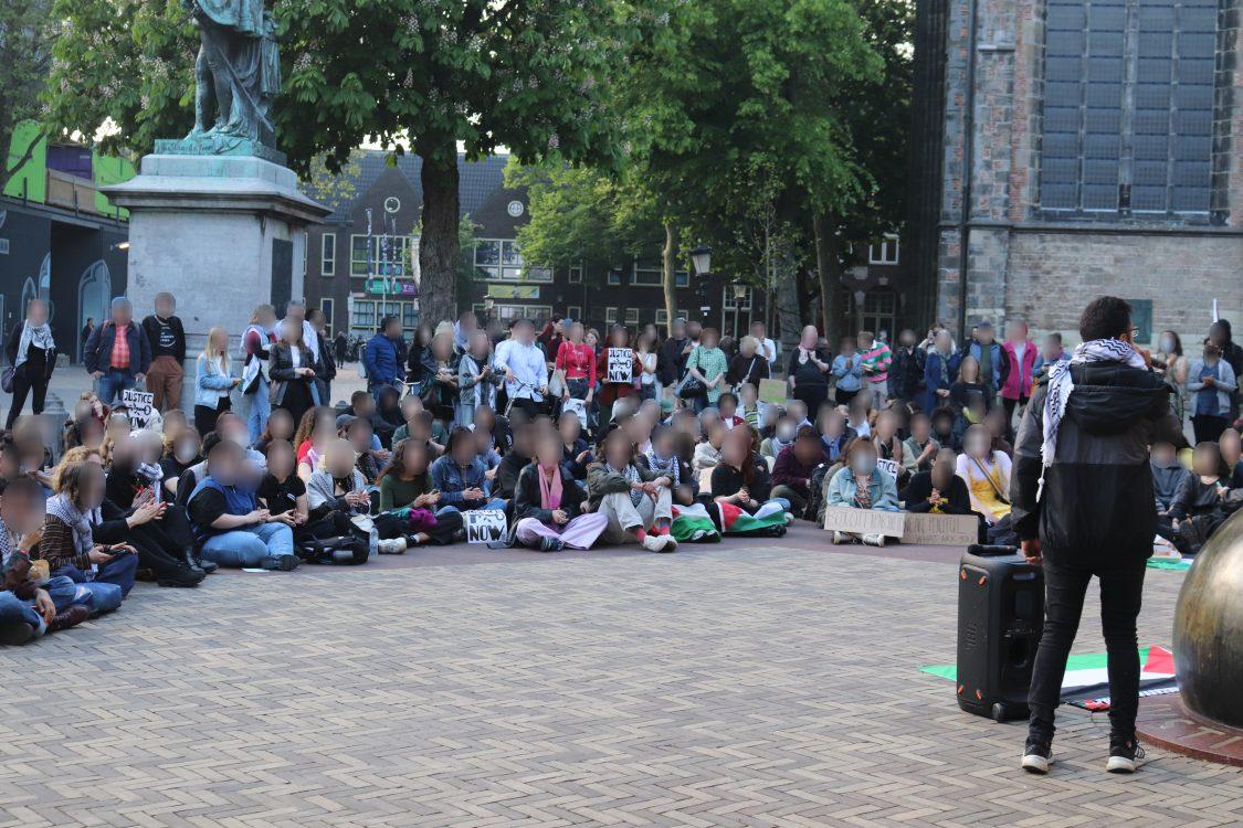 Een groep van honderden mensen zit en staat buiten in een halve cirkel op een plein. Velen dragen gezichtsmaskers. Ze dragen borden, spandoeken en Palestijnse vlaggen. Een persoon met een keffiyeh om, staand aan de rand van de menigte, spreekt de groep toe met microfoon in de hand en aan diens voeten een luidspreker. Naast Hij houdt een draagbare luidspreker vast en draagt een donker jasje en een geruite sjaal. Verschillende gebouwen en wat groene bomen zijn aanwezig op de achtergrond. Een tenminste zes meter hoog, klassiek standbeeld van een persoon op een voetstuk staat achter de menigte. Ook zijn linksachter bouwsteigers in beeld.