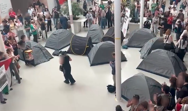 Een blik van bovenaf op een grote, felverlichte binnenruimte vol met mensen. Er staan ongeveer tien grijze kampeertenten rond de ruimte. Mensen staan en zitten rondom de tenten. Een persoon in het midden met een megafoon spreekt de mensen toe, een ander centraal persoon bekijkt diens camerabeelden. Een groep mensen klapt en juicht. Anderen houden borden, spandoeken, posters en Palestinavlaggen vast. Een persoon houdt een grote heuptrommel vast.