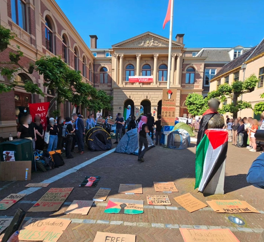 Op een zonnige binnenplaats omgeven door klassiek aandoende gebouwen, verschillende bomen en gehesen rode vlag staat een groep van tientallen mensen met borden en spandoeken naar het midden van de binnenplaats gericht. Er staat een stuk of vijf eenvoudige kampeertenten. Een standbeeld is in een Palestijnse vlag gewikkeld. Op de voorgrond liggen zo'n twintig kartonnen borden met symboliek en boodschappen ter ondersteuning van Palestina. Tussen twee bomen is een groot rood spandoek met witte letters gespannen. Veel mensen dragen gezichtsmaskers en keffiyeh.
