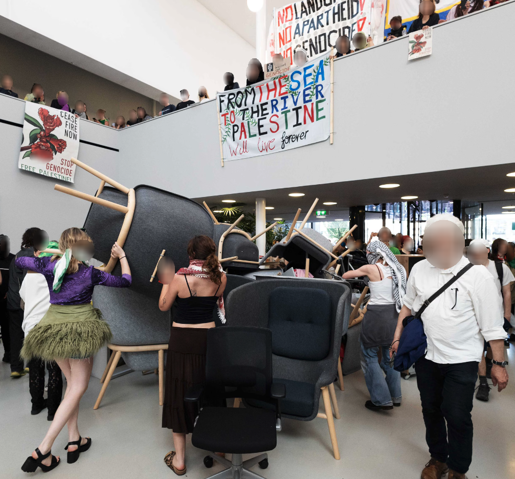 In een open, lichte binnenruimte met hoog plafond en meerdere open verdiepingen met balustrades stapelt een tiental mensen meubilair op elkaar. Velen van hen dragen keffiyeh. Over de balustrades van de eerste en tweede verdiepingen hangen grote spandoeken en posters naar beneden, met daarop teksten ter ondersteuning van Palestina. Tientallen mensen kijken vanaf de balustrades naar beneden. Op de rechtervoorgrond staan een persoon met onbedekt gezicht, neutrale kleding en een schoudertas die schuin uit het beeld kijkt.