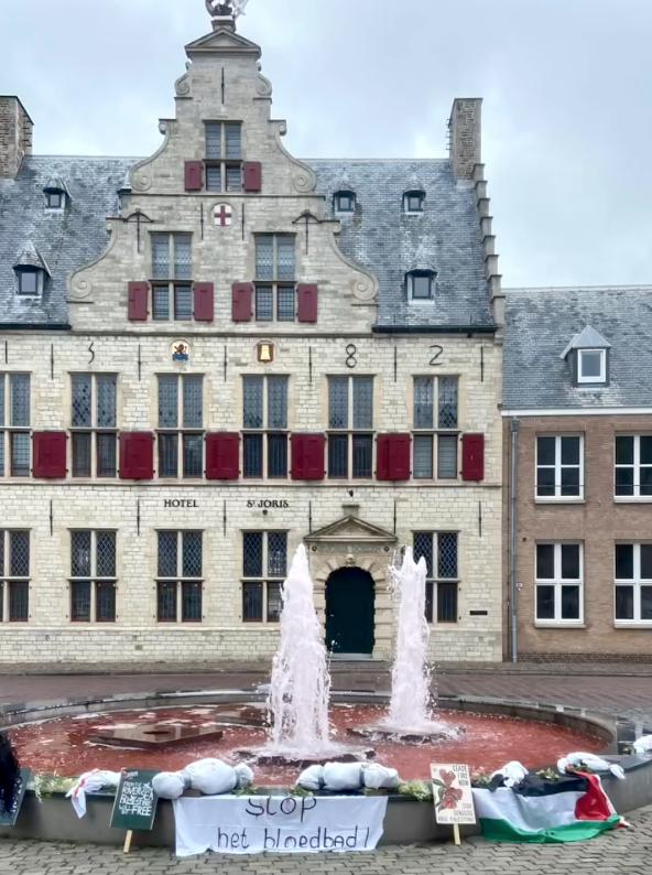 Op een plein staat een fontein. Het water in de fontein is rood gekleurd. Aan de rand van de fontein liggen beelden die bebloede kinderlijkjes in lijkzakken voorstellen. Ook liggen er Palestijnse vlaggen, posters en borden.