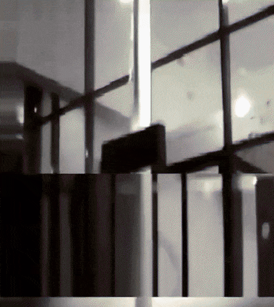 Aanzicht op een verlicht gebouw met een glazen voorgevel bij nacht. Twee donker gekleedde personen bespuiten de glazen voorgevel met rode verf.