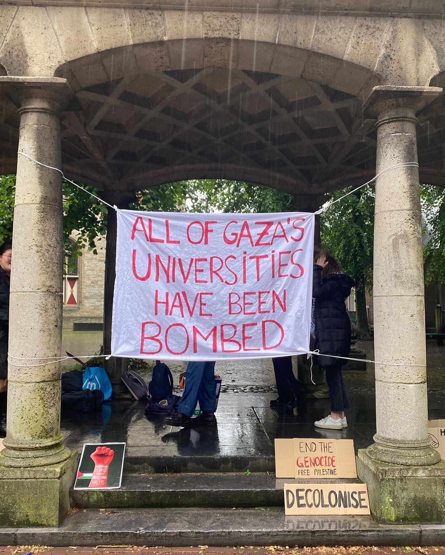 Een stenen overkapping in de regen. Tussen twee zuilen hangt een spandoek gespannen met de tekst (in het Engels): 'Alle van Gaza's universiteiten zijn gebombardeerd'. Bij de opgang staan borden en posters, waaronder met de teksten 'Dekoloniseer', 'Beëindig de genocide, vrij Palestina', en een poster met een rode, geheven vuist erop.