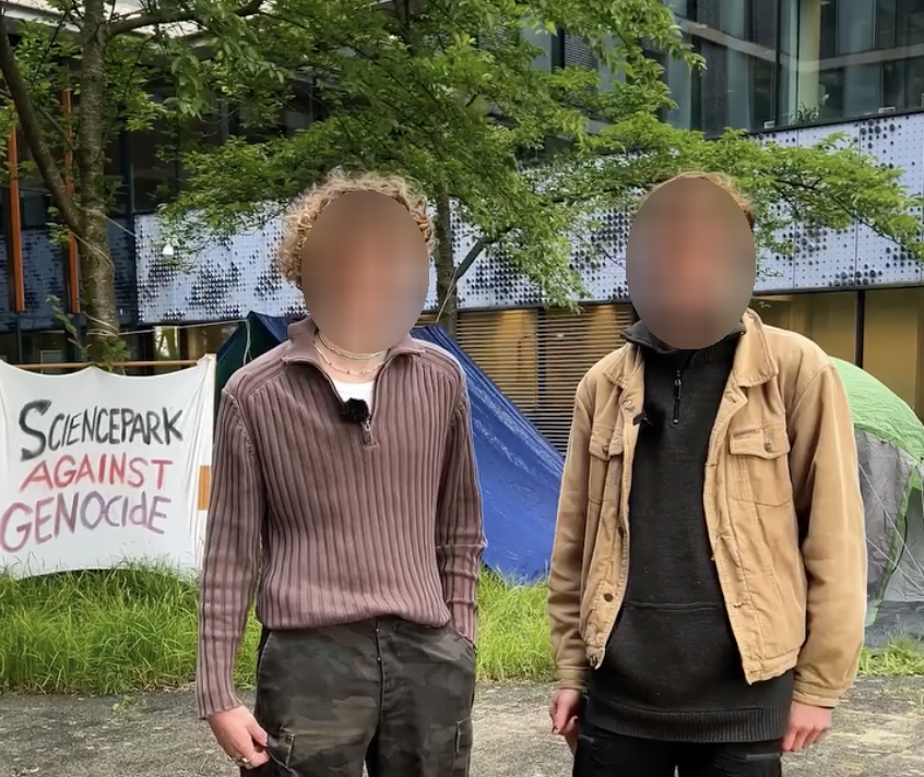 Twee personen in neutrale, loszittende kleding staan buiten voor een gebouw met houten, metaal en glazen bekleding en daarvoor verschillende bomen. De personen glimlachen en hun armen handen rustig langs hun lichaam. Op de achtergrond staan een blauwe en een groen-grijze kampeertent, met een spandoek met daarop de boodschap (in het Engels): 'Wetenschapspark tegen genocide'.