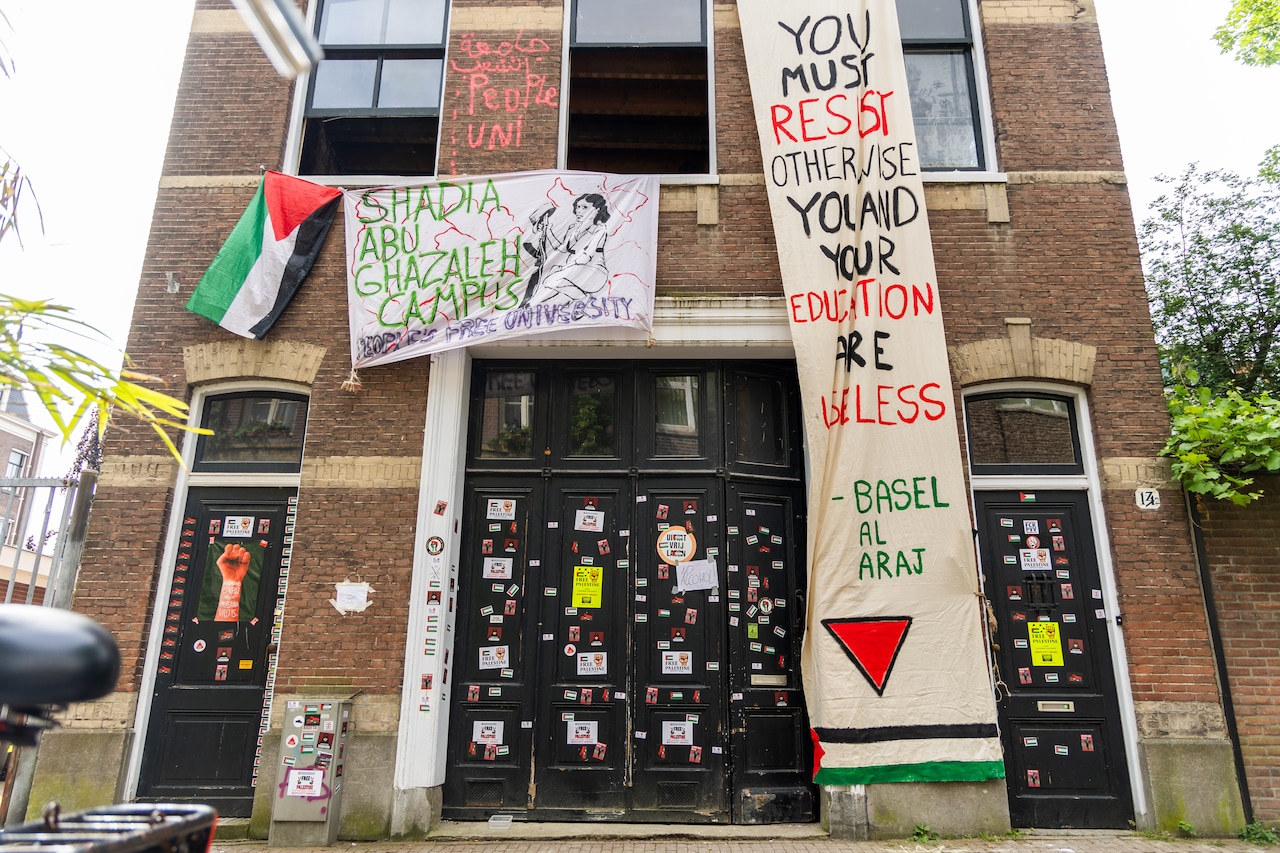 Een frontaal zicht op een bakstenengebouw van meerdere verdiepingen, met hoge donkeredeuren en ramen. Het is een bewolkte dag. Aan de rechterzijde zijn over een muur heen bomen te zien. Op de deuren op de begane grond zitten posters, flyers en stickers ter ondersteuning van Palestina. Een groot handgeschilderd spandoek vanaf de eerste verdieping toont een persoon met een wapen, en daarnaast in groene letters (onder meer) de tekst 'Shadia Abu Ghazaleh Campus'. Vanaf de tweede verdieping tot aan de straat is een langere, smallere banner met daarop de tekst (in het Engels), in zwarte, rode en groene leters: '... je moet je verzetten anders zijn jij en je opleiding waardeloos, Basel al Araj', met daaronder een rode driehoek en een Palestijnse vlag.