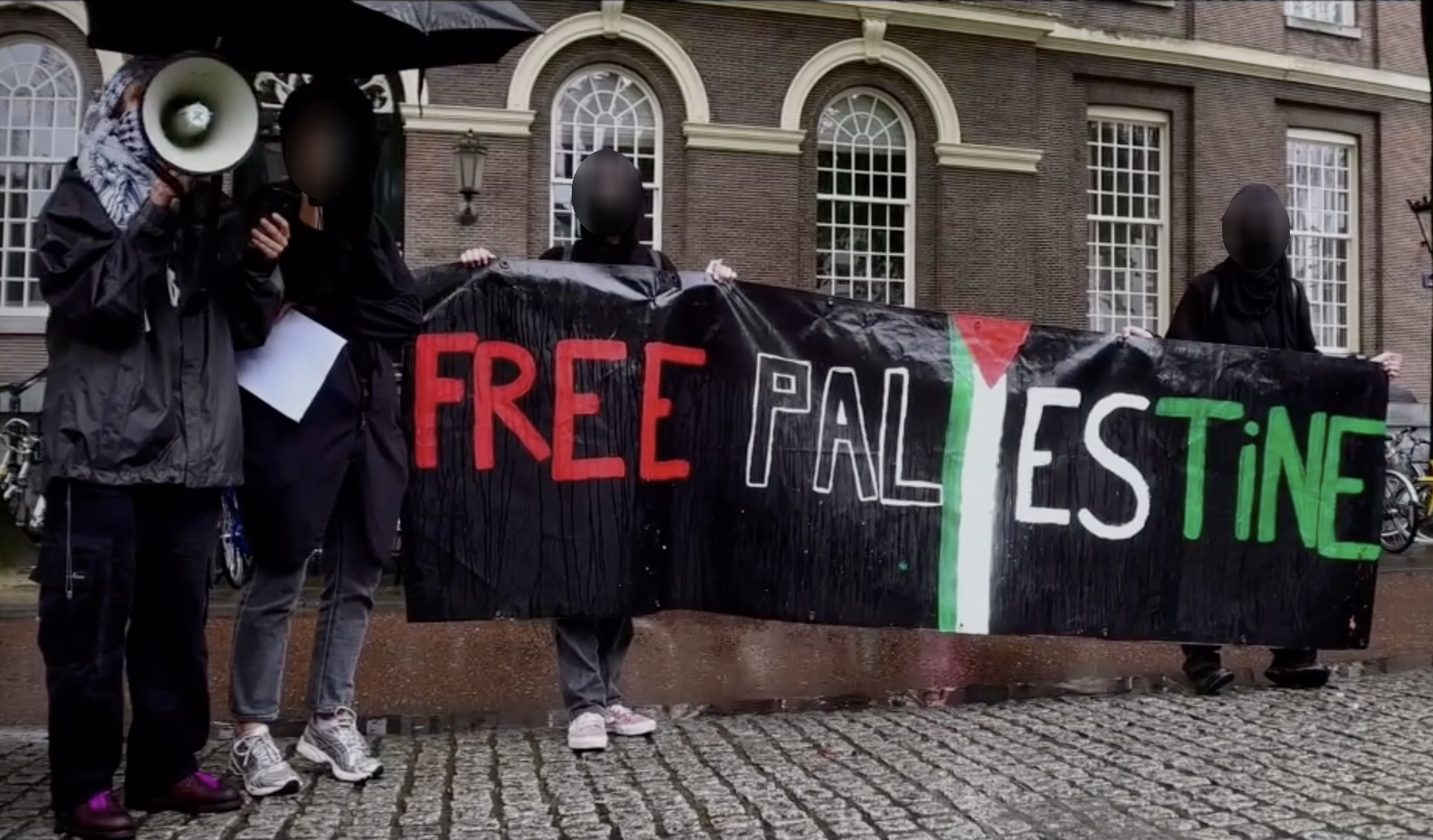 Vier mensen in voornamelijk donkere kleding staan in de stromende regen op de kinderkoppenstraat voor een fietsstalplaats, voor een historisch bakstenen gebouw met hoge boogramen. Aan de linkerzijde staan twee personen onder een zwarte paraplu: één met een megafoon en een apparaat in de hand, de ander houdt een leeg wit blad en de paraplu vast. De andere twee personen houden een zwarte banner vast met daarop in rode, witte en groene letters de tekst (in het Engels) 'Bevrijd Palestina', samen met een geroteerde Palestijnse vlag.