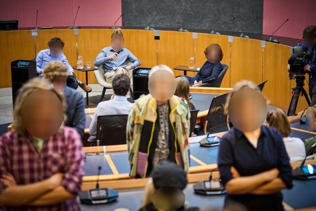 In een formeel uitziende vergaderzaal, met een houtpanelen spreekgedeelte en individuele zetels met microfoons, is een handvol mensen aanwezig. In het midden, op lagere hoogte, zitten twee personen in lichtblauwe overhemden zonder das of jasje, met één van hen een microfoon in de hand. Naast hen zit een persoon in donkere kleding, die een blik werpt richting de camera en de hoger gelegen zetelgedeeltes. Op die hogere zetels kijken een aantal zittende mensen recht naar voren, terwijl een ander aantal zich staande, met de rug naar het centrum, demonstratief heeft opgedraaid.