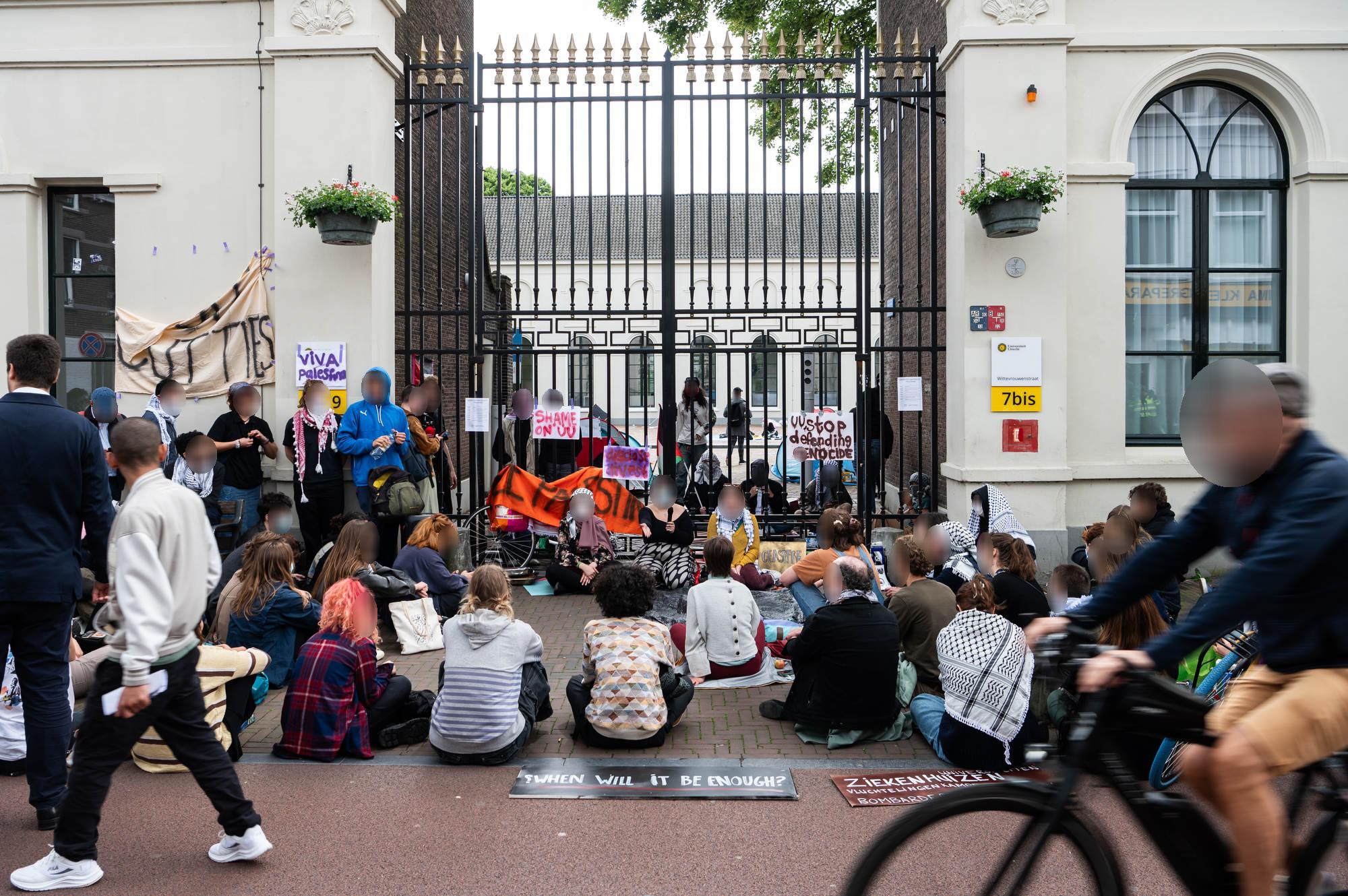 Op straat, voor een wit gebouw met hoge ramen en een metershoog hek met donkere spijlen en goude punten, zitten en staan tientallen mensen, op de rug gezien. Zij hebben bordjes en spandoeken bij zicht ter ondersteuning van Palestina, met teksten zoals (in het Engels) 'Schaam je UU',en 'UU stop met het verdedigen van genocide'. Tegen het het geleund zit een drietal mensen, waarvan één met een microfoon de groep toespreekt. Aan de andere zijde, achter het hek, zitten verschillende mensen die keffiyeh dragen naast een aantal kleurrijke kampeertenten. Op de voorgrond passeren twee voetgangers en een fietser, die toekijken.